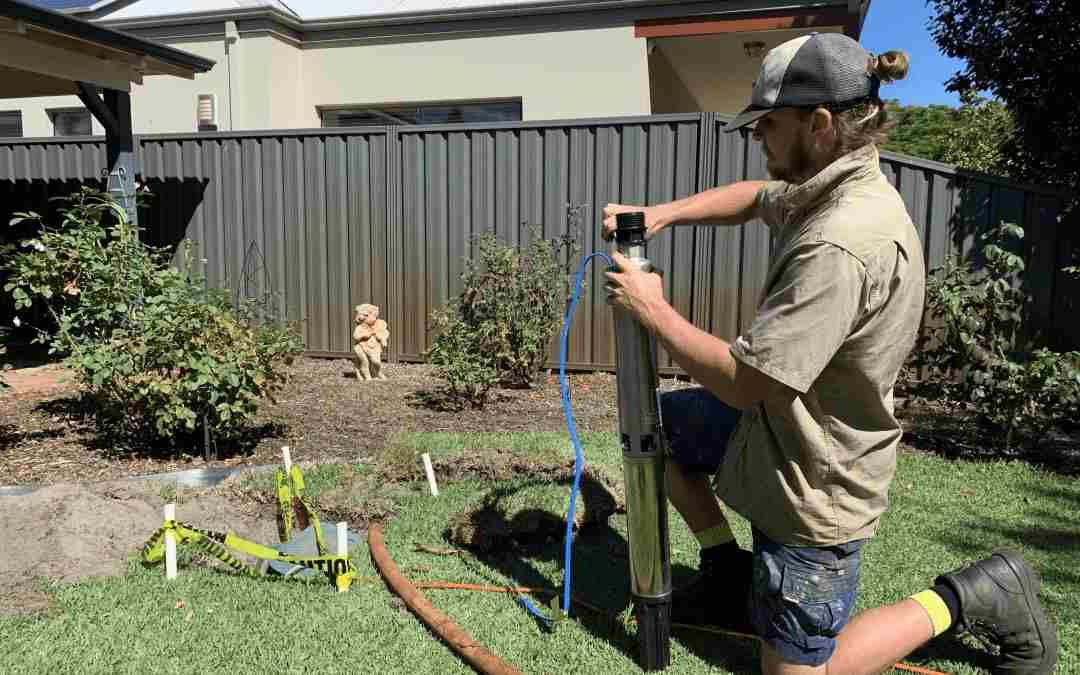 Technician kneeling on a lawn lifting a submersible bore pump from its borehole next to caution tape.